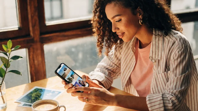 Una donna sorride mentre compone un collage di foto sul suo cellulare, con foto stampate e caffè sul tavolo.